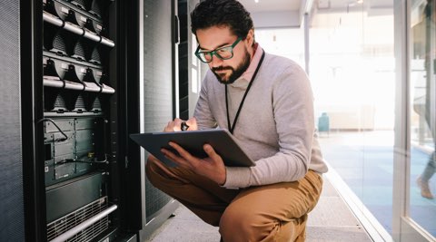 Male IT specialist in a server work, kneeling while checking his tablet.
